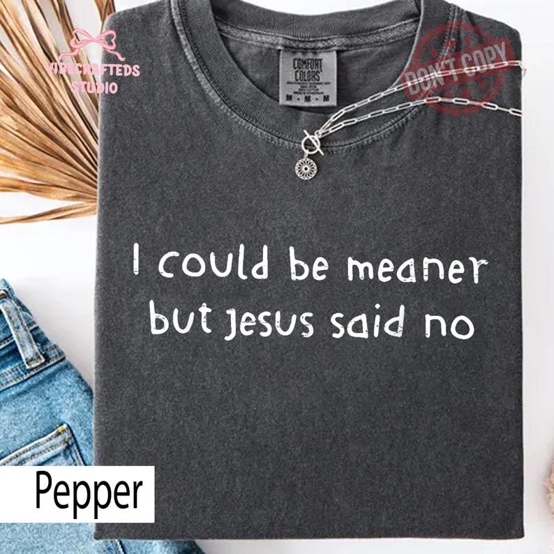 May include: A dark gray t-shirt with the text "I could be meaner but Jesus said no" in white. The shirt has a crew neck and short sleeves. A silver necklace with a pendant is draped on the shirt. The word "Pepper" is at the bottom.