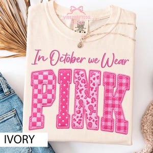 May include: An ivory t-shirt featuring the text In October we Wear PINK in a pink script and block font. The letters are filled with various pink patterns including checkers, polka dots, leopard print with ribbons, and a plaid design.