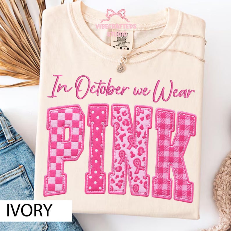 May include: An ivory t-shirt featuring the text In October we Wear PINK in a pink script and block font. The letters are filled with various pink patterns including checkers, polka dots, leopard print with ribbons, and a plaid design.
