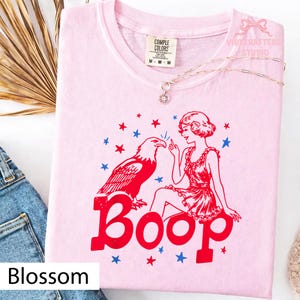 May include: A pale pink t-shirt featuring a red eagle, a woman in a red dress, and the word "Boop" in red. Blue and red stars are scattered around the design. The shirt has a crew neck and short sleeves.