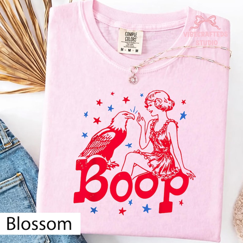 May include: A pale pink t-shirt featuring a red eagle, a woman in a red dress, and the word "Boop" in red. Blue and red stars are scattered around the design. The shirt has a crew neck and short sleeves.