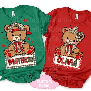 May include: Two t-shirts, one green and one red, each featuring a cartoon teddy bear holding a heart. The green shirt has the name "MATHEW" on a sign, while the red shirt has "OLIVIA". Both shirts have a Valentine's Day theme.