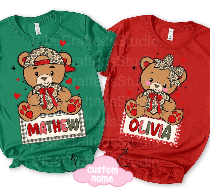 May include: Two t-shirts, one green and one red, each featuring a cartoon teddy bear holding a heart. The green shirt has the name "MATHEW" on a sign, while the red shirt has "OLIVIA". Both shirts have a Valentine's Day theme.