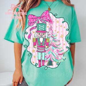 May include: A light teal t-shirt with a festive design. It features a nutcracker soldier, a Christmas tree, and a large pink bow. The nutcracker is in pink, green, and gold. The background has pink and white stripes. The design is inside a black outlined frame.
