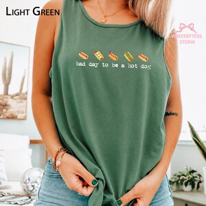 May include: A light green sleeveless tank top featuring a graphic of five hot dogs arranged in a horizontal row above the white text that reads, "bad day to be a hot dog." The shirt has a round neckline and a relaxed fit design.