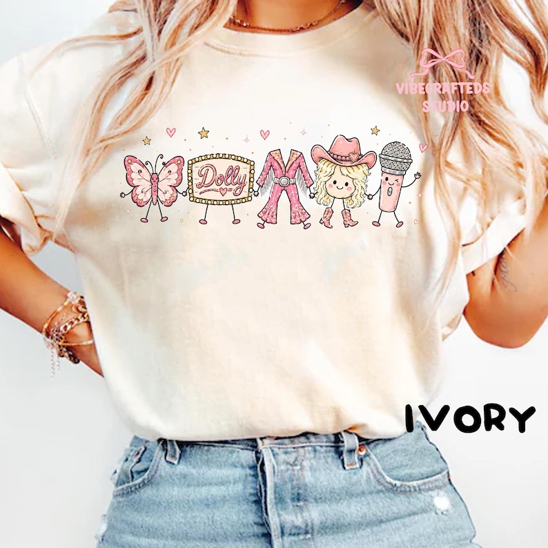 May include: An ivory t-shirt featuring a graphic print of a pink butterfly, a marquee sign with the text Dolly, a pink fringe outfit, a blonde character wearing a pink cowboy hat, and a microphone, all holding hands with small hearts and stars.