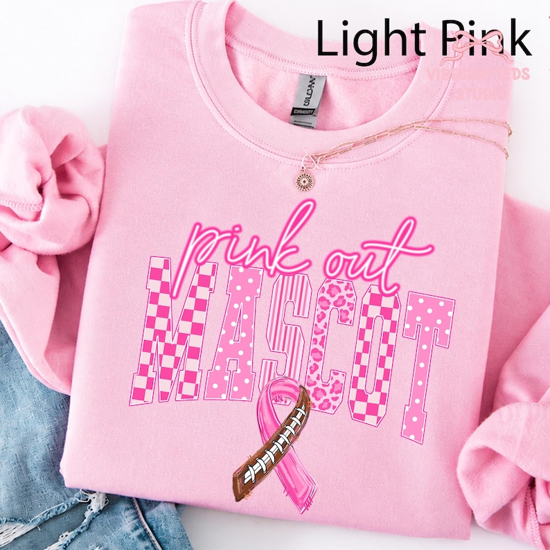 May include: A light pink crewneck sweatshirt featuring the text "pink out MASCOT" in a mix of checkered, polka dot, and leopard print patterns. A pink awareness ribbon designed with football laces is centered below the text.