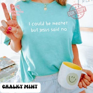 May include: A light blue t-shirt with the text "I could be meaner but jesus said no." The shirt is paired with white shorts and a white mug with a yellow interior and a smiley face.