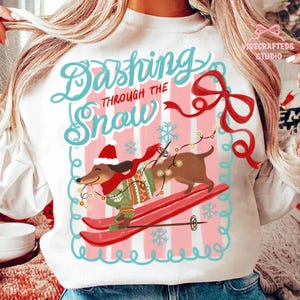 May include: White sweatshirt with a festive design. The graphic features a dachshund dog skiing, wearing a hat, scarf, and sweater, with the words "Dashing Through The Snow". The design includes snowflakes and a red bow.