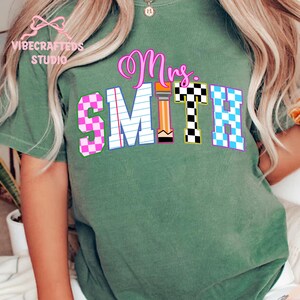 May include: Green t-shirt with the name "Mrs. Smith" in a colorful, playful design. The letters are in various patterns, including pink and blue checkered, and a pencil graphic. The shirt has the "VIBECRAFTED STUDIO" logo.