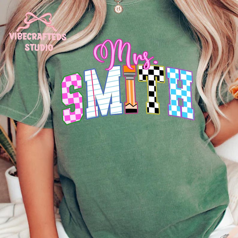 May include: Green t-shirt with the name "Mrs. Smith" in a colorful, playful design. The letters are in various patterns, including pink and blue checkered, and a pencil graphic. The shirt has the "VIBECRAFTED STUDIO" logo.