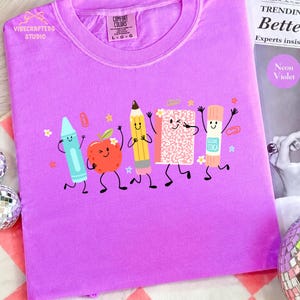 Puede incluir: Una camiseta de color violeta ne&oacute;n con un estampado gr&aacute;fico de cinco &uacute;tiles escolares antropom&oacute;rficos, incluyendo un cray&oacute;n azul, una manzana roja, un l&aacute;piz amarillo, un cuaderno rosa moteado y un pegamento en barra, con caras sonrientes.