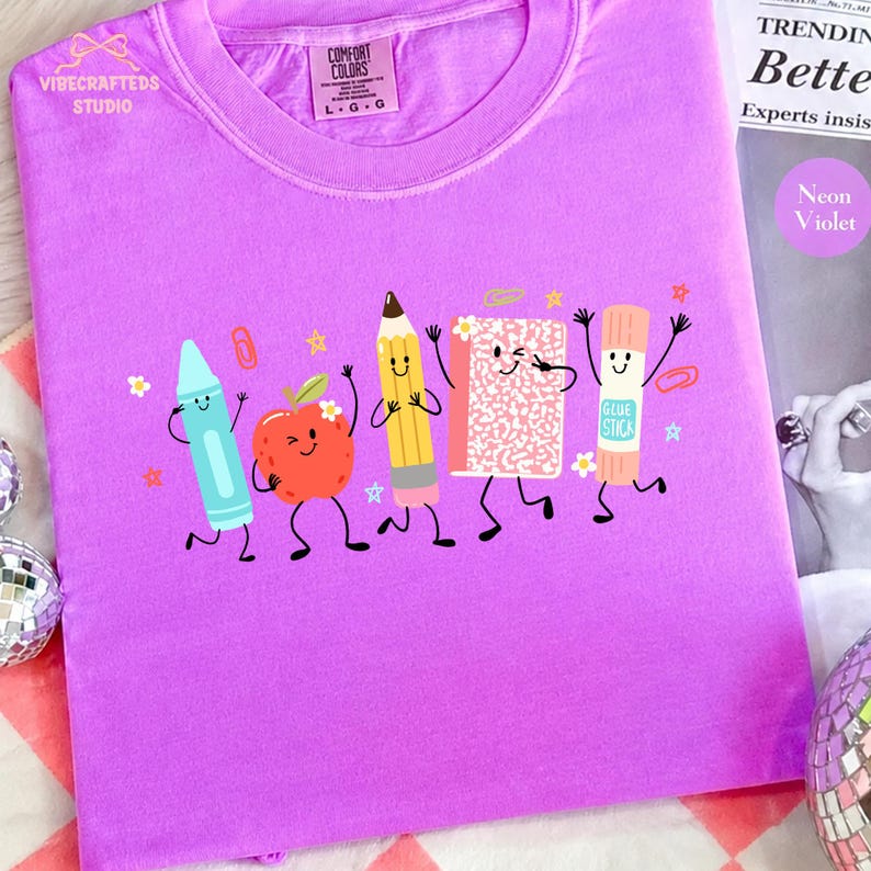 Puede incluir: Una camiseta de color violeta ne&oacute;n con un estampado gr&aacute;fico de cinco &uacute;tiles escolares antropom&oacute;rficos, incluyendo un cray&oacute;n azul, una manzana roja, un l&aacute;piz amarillo, un cuaderno rosa moteado y un pegamento en barra, con caras sonrientes.