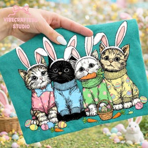 May include: Teal graphic tee featuring four kittens dressed for Easter. Each kitten wears bunny ears and a sweater. One kitten has a blue bow tie, another holds a carrot, and all are surrounded by Easter eggs and carrots. The text "VIBECRAFTEDS STUDIO" is visible.