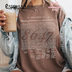Puede incluir: Una sudadera marrón con el texto de la Declaración de Independencia en blanco. La palabra "SOY" se muestra en una fuente grande y estilizada. La sudadera tiene cuello redondo y mangas largas.