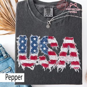 Pu&ograve; includere: Una maglietta grigio scuro con la scritta "USA" in un design a bandiera americana usurata. Le lettere sono realizzate in denim e tessuto rosso e bianco. La maglietta ha un girocollo e maniche corte.