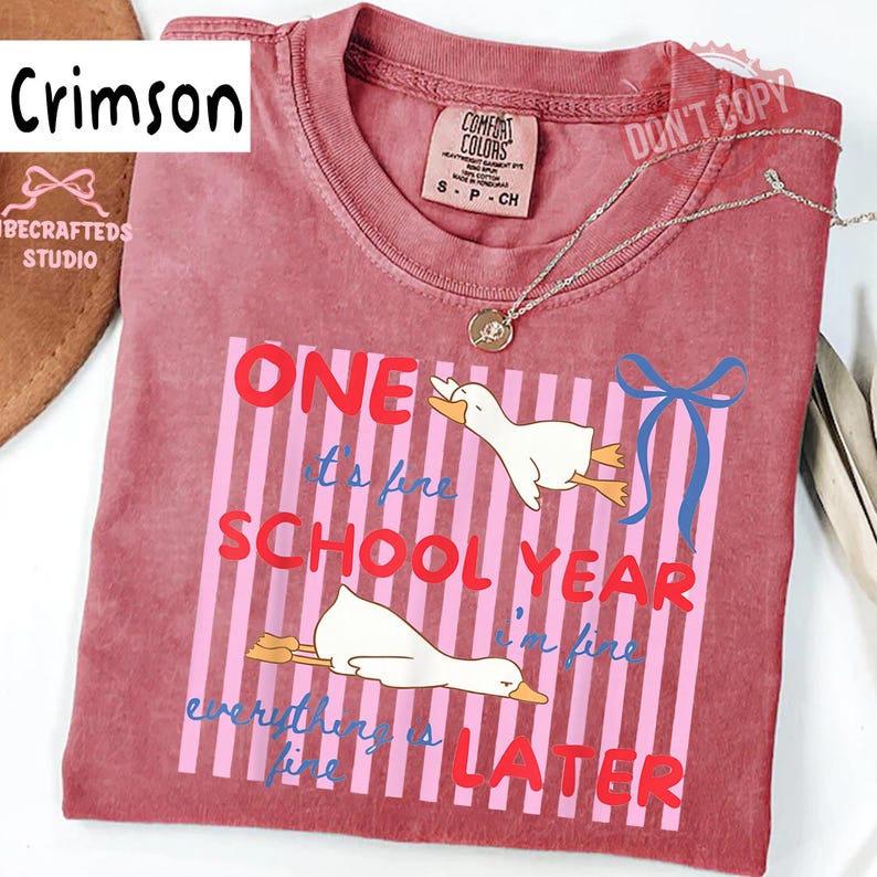 May include: Crimson-colored t-shirt with the text "ONE SCHOOL YEAR LATER" and two cartoon ducks. The shirt also has the text "it's fine, I'm fine, everything is fine." A silver necklace and a tag are visible.