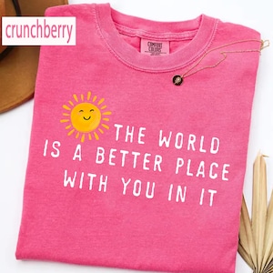 May include: A bright pink short-sleeved t-shirt featuring a yellow smiling sun graphic and the white printed text, "THE WORLD IS A BETTER PLACE WITH YOU IN IT." The shirt has a crew neckline and a Comfort Colors brand label on the inner collar.