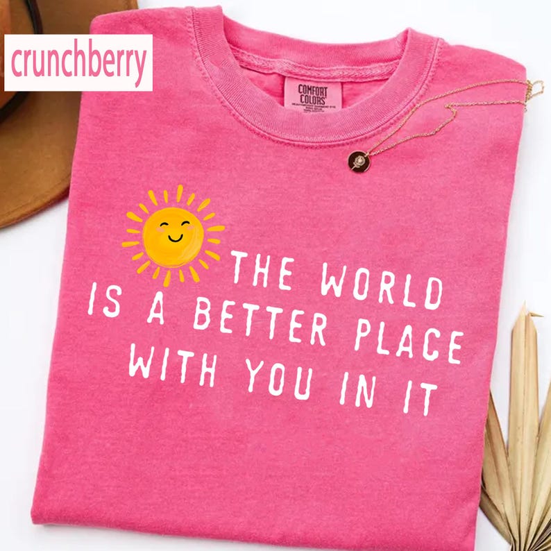 May include: A bright pink short-sleeved t-shirt featuring a yellow smiling sun graphic and the white printed text, "THE WORLD IS A BETTER PLACE WITH YOU IN IT." The shirt has a crew neckline and a Comfort Colors brand label on the inner collar.