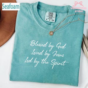 May include: Seafoam green t-shirt with the text "Blessed by God, Loved by Jesus, Led by the Spirit" in white script. The shirt has a crew neck and short sleeves. A gold-coloured necklace with a pendant is draped on the shirt.