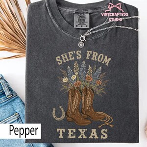 May include: Dark gray Comfort Colors t-shirt with the text "She's From Texas". The shirt features a graphic of brown cowboy boots, flowers, and a horseshoe. A silver necklace with a pendant is draped on the shirt.
