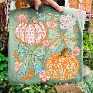 May include: A green t-shirt featuring a fall-themed design. The design includes two pumpkins, two bows, and floral accents in shades of orange, pink, and blue. The pumpkins have floral patterns, and the bows have glitter accents.