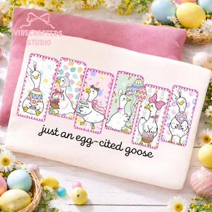 May include: A cream-colored t-shirt featuring six cartoon geese, each with Easter-themed decorations and holding Easter eggs or baskets. The text "just an egg-cited goose" is printed below the geese. The shirt is on a pink pillow.