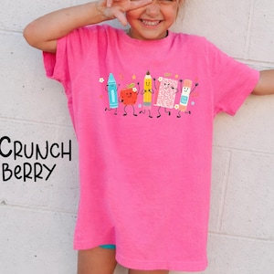 Puede incluir: Una camiseta de color rosa brillante de manga corta con un estampado gr&aacute;fico de cinco &uacute;tiles escolares antropom&oacute;rficos que incluyen un cray&oacute;n azul, una manzana roja, un l&aacute;piz amarillo, un cuaderno rosa y un pegamento, con el texto Crunch Berry.