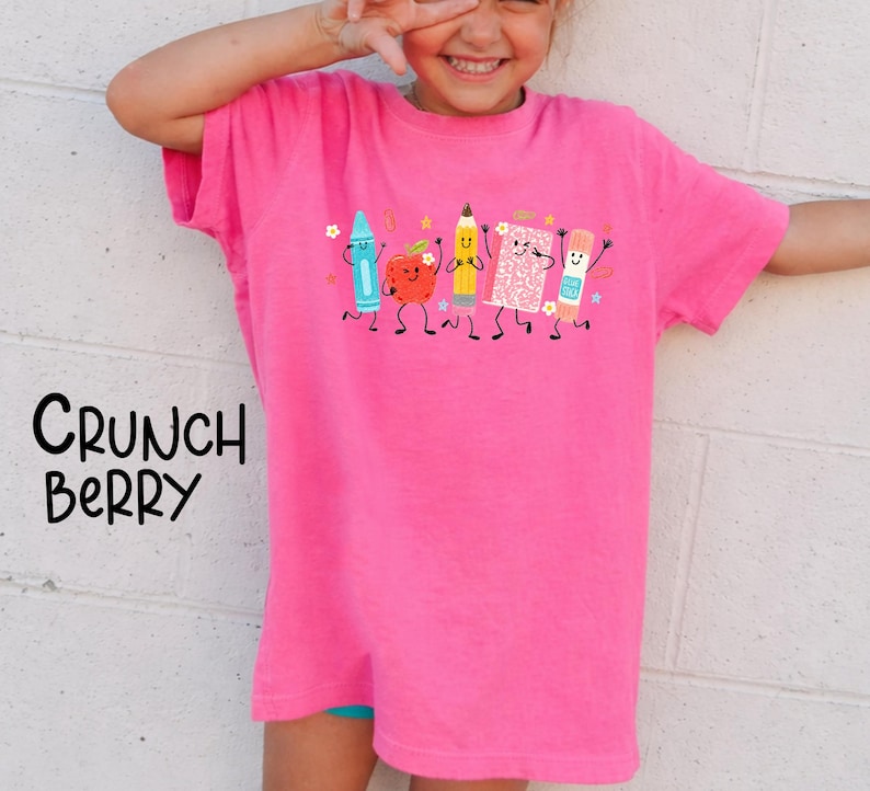 Puede incluir: Una camiseta de color rosa brillante de manga corta con un estampado gr&aacute;fico de cinco &uacute;tiles escolares antropom&oacute;rficos que incluyen un cray&oacute;n azul, una manzana roja, un l&aacute;piz amarillo, un cuaderno rosa y un pegamento, con el texto Crunch Berry.