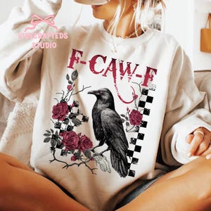 May include: Cream-colored sweatshirt featuring a black raven perched on a branch with red roses and the text "F-CAW-F" in red. A black and white checkered pattern is on the right side of the raven. The sweatshirt has long sleeves.