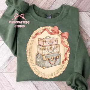 May include: Green sweatshirt featuring a vintage-style illustration of three stacked suitcases within an oval frame, accented with a pink ribbon. The text "VIBECRAFTED STUDIO" is printed on the upper left sleeve.