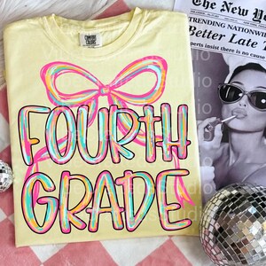 Pode incluir: Camiseta amarelo claro com as palavras "FOURTH GRADE" em uma fonte colorida e delineada, e um gr&aacute;fico de la&ccedil;o. A camiseta &eacute; feita de um material macio. Um jornal e uma bola de discoteca est&atilde;o no fundo.