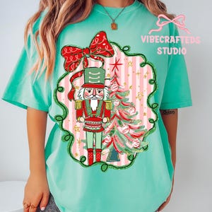 May include: A mint green t-shirt featuring a Christmas-themed graphic. The graphic includes a nutcracker soldier, a Christmas tree, and a red bow. The design is set against a pink and white striped background, with the text "VIBECRAFTED STUDIO".