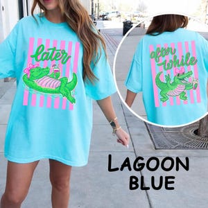 Puede incluir: Camiseta azul laguna con un gráfico de un caimán verde con un lazo rosa y una bebida. La parte delantera de la camiseta dice "later" y la trasera "after while". Rayas verticales rosas están detrás del caimán.