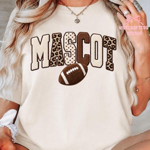 May include: A cream-colored t-shirt featuring the word MASCOT in large, textured block letters with alternating leopard print and solid brown patterns. A brown American football graphic is positioned below the text. The text VIBECRAFTED STUDIO appears in the upper right corner.