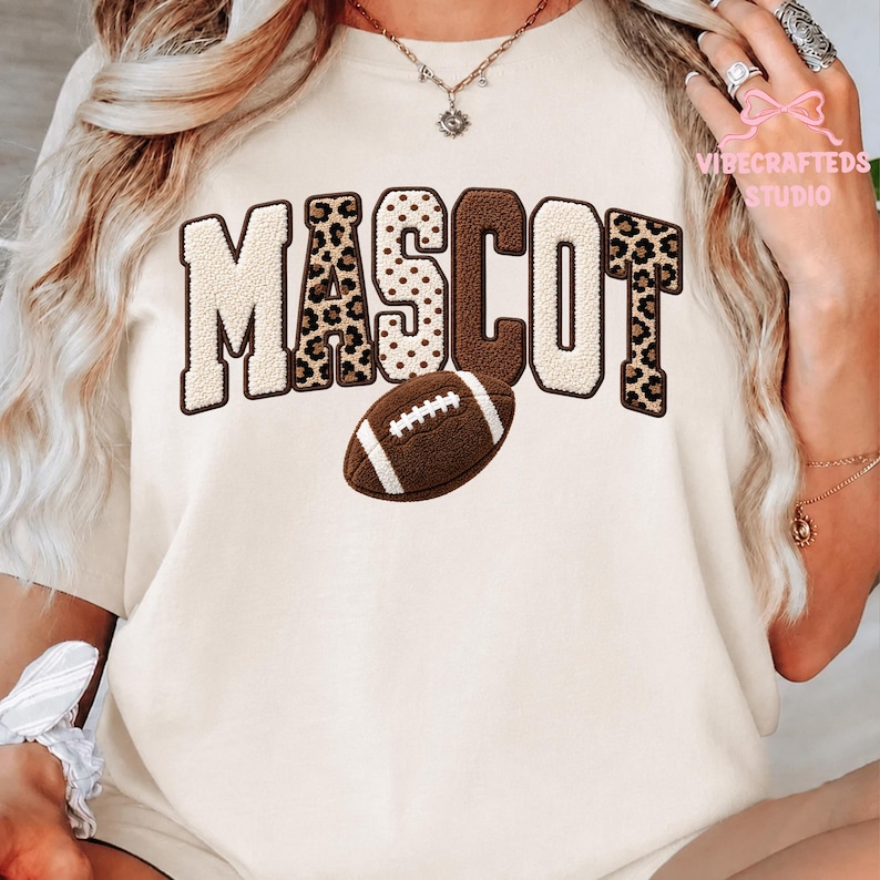 May include: A cream-colored t-shirt featuring the word MASCOT in large, textured block letters with alternating leopard print and solid brown patterns. A brown American football graphic is positioned below the text. The text VIBECRAFTED STUDIO appears in the upper right corner.