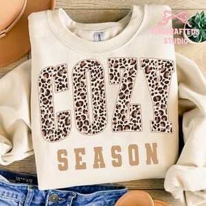 May include: Cream-colored sweatshirt with the words "COZY SEASON" printed on the front. The word "COZY" is in a leopard print pattern, and "SEASON" is in a solid brown color. The sweatshirt is folded on a wooden surface.
