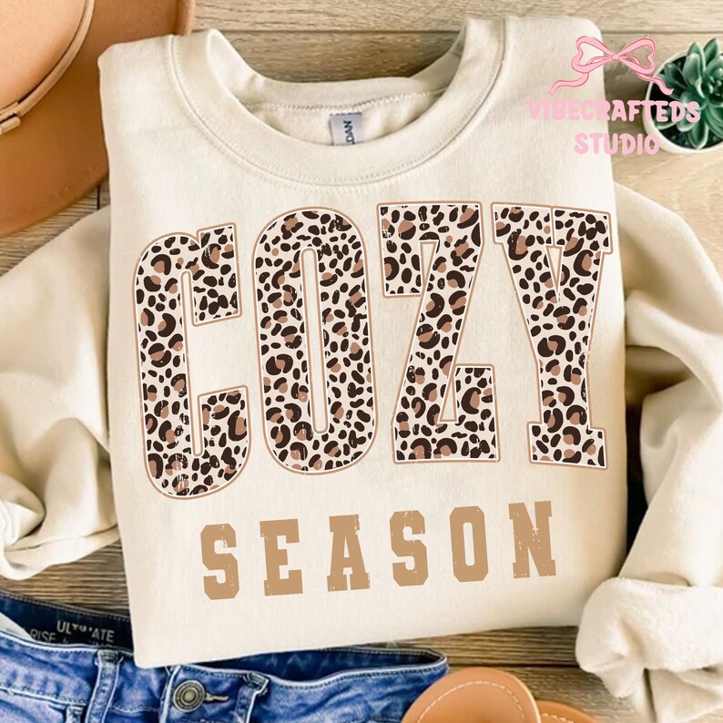 May include: Cream-colored sweatshirt with the words "COZY SEASON" printed on the front. The word "COZY" is in a leopard print pattern, and "SEASON" is in a solid brown color. The sweatshirt is folded on a wooden surface.