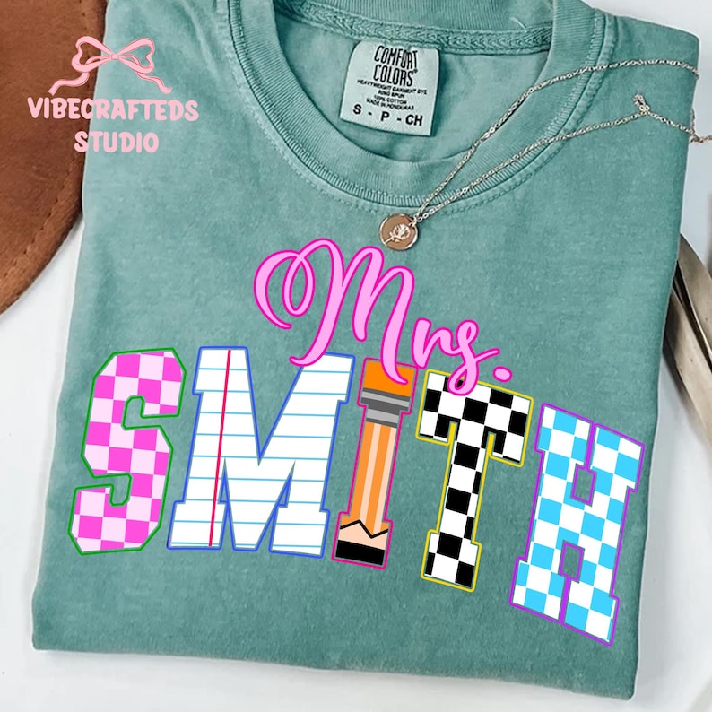 May include: A teal Comfort Colors t-shirt with the name "Mrs. SMITH" in a colorful, playful font. The letters are decorated with patterns like pink checkerboard, lined paper, and a pencil design. A silver necklace with a pendant is draped across the shirt.