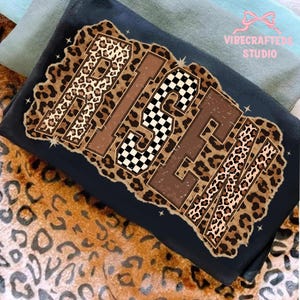 May include: Black t-shirt featuring the word "RISE" in a distressed leopard print design. The letter "S" has a checkered pattern. The text "VIBECRAFTED STUDIO" is in the upper right corner. The shirt is folded on a leopard print fabric.