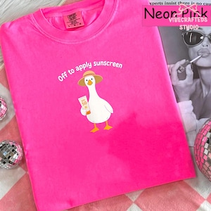 Pode incluir: Camiseta rosa brilhante com a frase "Off to apply sunscreen". Uma ilustração de um ganso de desenho animado usando chapéu e segurando protetor solar é impressa na frente. Marca Comfort Colors.