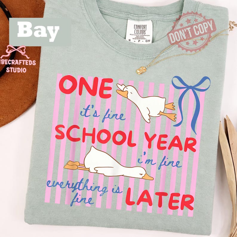 May include: A light green t-shirt with a graphic design. The design features two cartoon ducks, pink and red text that reads "ONE SCHOOL YEAR LATER", and the phrase "it's fine, everything is fine, I'm fine".