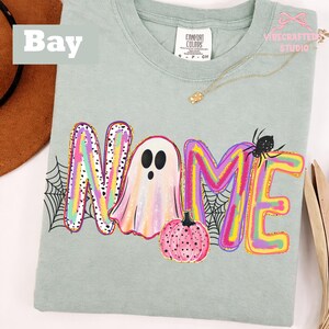 May include: A light sage green t-shirt featuring a graphic print of the word NAME, where the letter A is replaced by a white ghost. The letters are filled with colorful brushstrokes, and the design includes a pink pumpkin and a black spider.