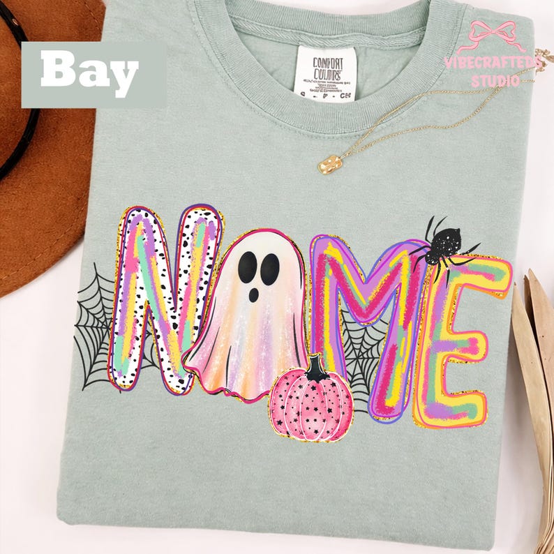 May include: A light sage green t-shirt featuring a graphic print of the word NAME, where the letter A is replaced by a white ghost. The letters are filled with colorful brushstrokes, and the design includes a pink pumpkin and a black spider.