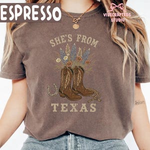 May include: Espresso-colored t-shirt with a graphic design featuring cowboy boots, flowers, and the text "She's From Texas." A horseshoe and rope are also included in the design. The shirt has a vintage, worn look.