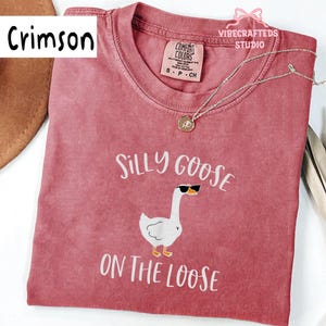 May include: Crimson-colored t-shirt with the text "Silly Goose On The Loose" and a graphic of a goose wearing sunglasses. The shirt has a crew neck and a small tag at the neckline. A silver necklace with a pendant is draped across the shirt.