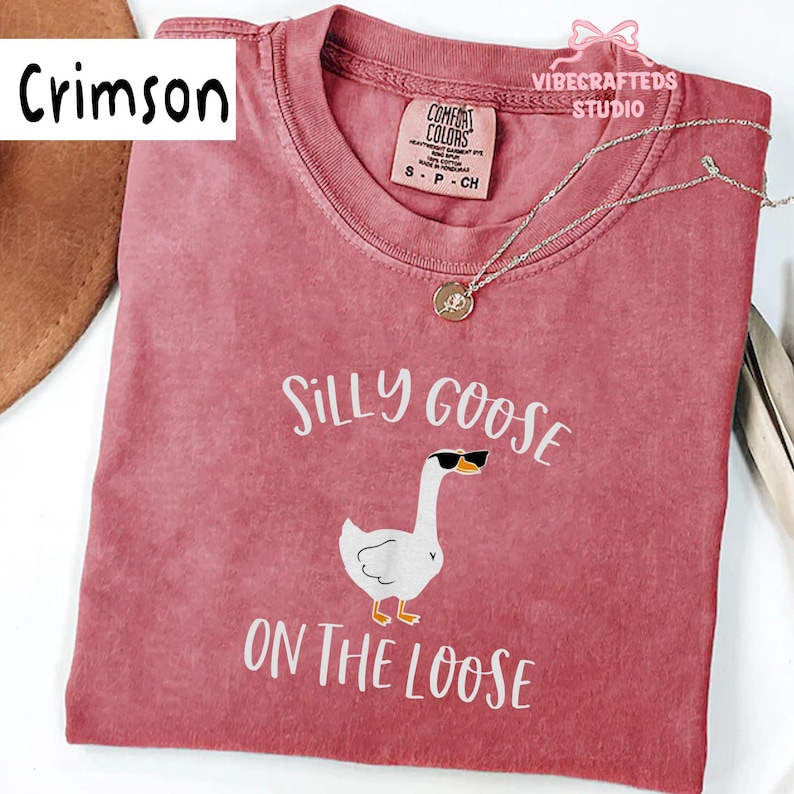 May include: Crimson-colored t-shirt with the text "Silly Goose On The Loose" and a graphic of a goose wearing sunglasses. The shirt has a crew neck and a small tag at the neckline. A silver necklace with a pendant is draped across the shirt.