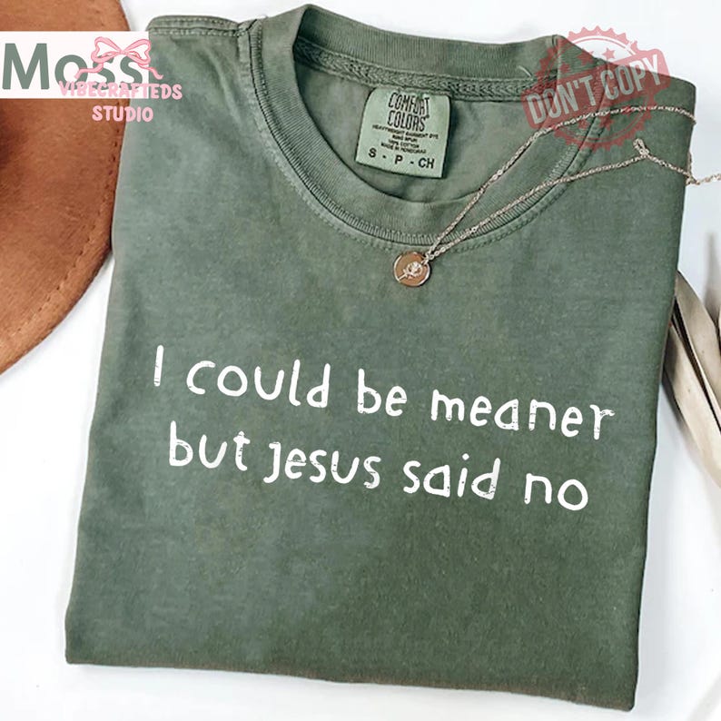 May include: An olive green t-shirt with the text "I could be meaner but Jesus said no" in white. The shirt has a crew neck and a small tag that says "Comfort Colors". A silver necklace with a pendant is on the shirt.