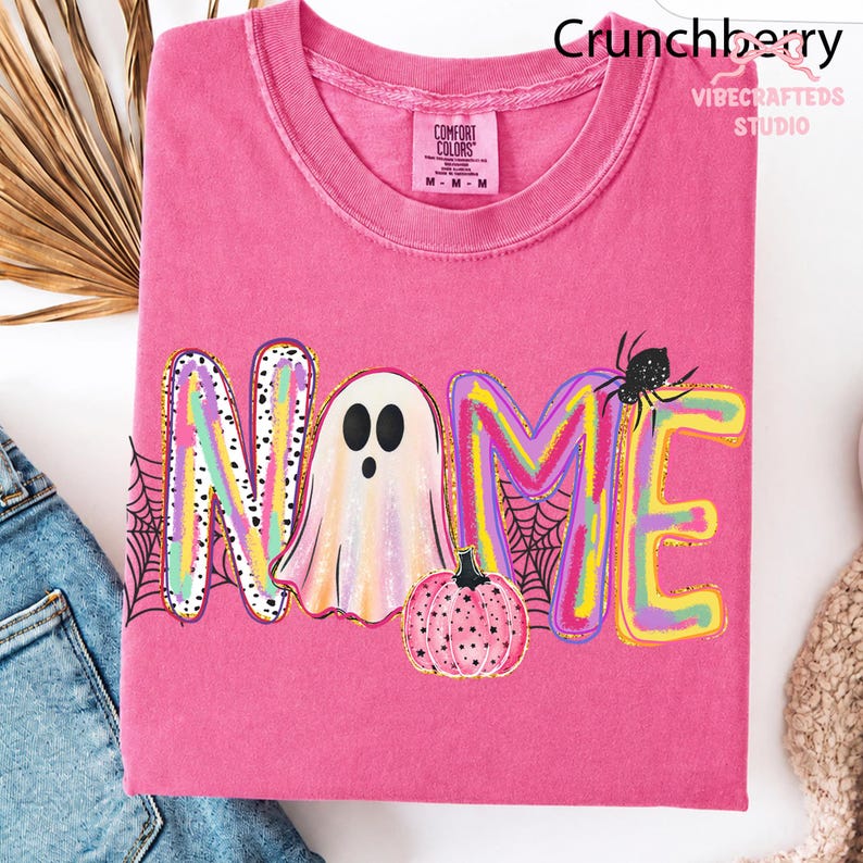 May include: A bright pink short-sleeve t-shirt featuring a colorful graphic with the word NAME, where the letter A is replaced by a ghost. The design includes a pink pumpkin, a black spider, and spiderwebs. The shirt has a Comfort Colors label.