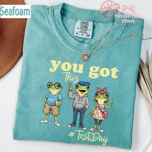 Puede incluir: Camiseta verde mar con la frase "you got this" y tres ranas de dibujos animados. Las ranas llevan gafas, un sombrero y un vestido floral. El texto "#Test Day" está debajo. Se ve una etiqueta Comfort Colors.